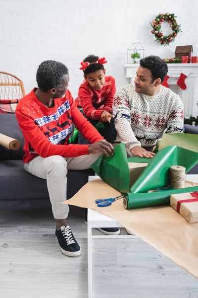 African american boy in reindeer horns headband pointing at table with gift boxes and wrapping paper near dad and grandpa — Stock Photo
