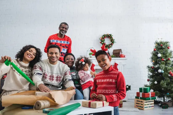 Familia afroamericana feliz mirando a la cámara mientras empaca regalos en la sala de estar con decoración de Navidad - foto de stock