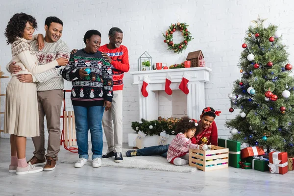Glücklich afrikanisch-amerikanische Familie sieht Bruder und Schwester spielen in der Nähe von Weihnachtsbaum im Wohnzimmer — Stockfoto