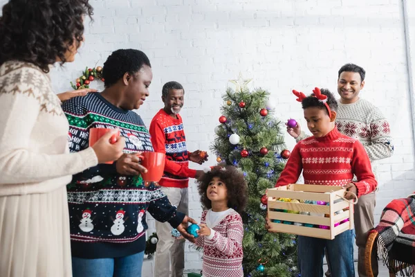 Afrikanisch-amerikanischer Junge in Rentierhörnern Stirnband hält Holzkiste mit dekorativen Kugeln in der Nähe von Familie und Weihnachtsbaum — Stockfoto