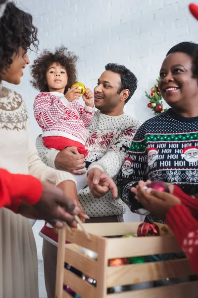 Sorrindo homem segurando filha e apontando para caixa de madeira com bolas de Natal perto de família afro-americana turva — Fotografia de Stock