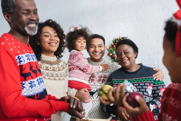 Joyeuse famille afro-américaine regardant un garçon flou tenant des boules de Noël — Photo de stock