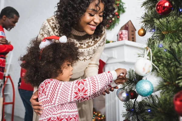 Afrikanerin mit Weihnachtsmütze schmückt Weihnachtsbaum in der Nähe von glücklicher Mutter — Stockfoto