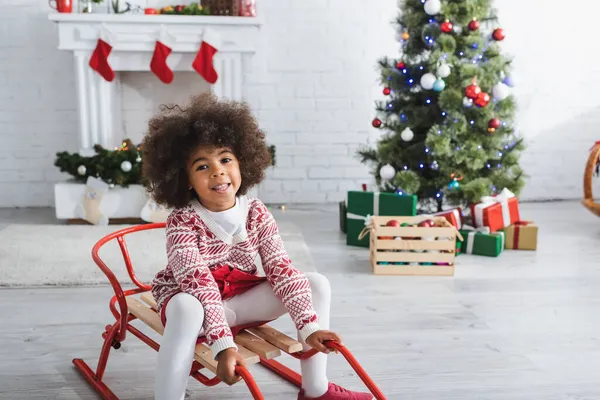 Fröhliche afrikanisch-amerikanische Kind sitzt auf Schlitten im Wohnzimmer mit dekoriertem Kamin und Weihnachtsbaum auf verschwommenem Hintergrund — Stockfoto