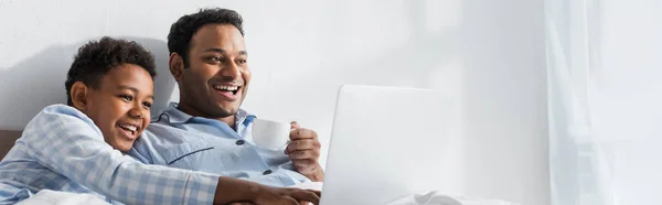 Laughing african american boy pointing at laptop while watching movie with dad in bedroom, banner — Stock Photo