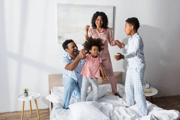 Excited african american family in pajamas holding hands while fooling around on bed — Stock Photo
