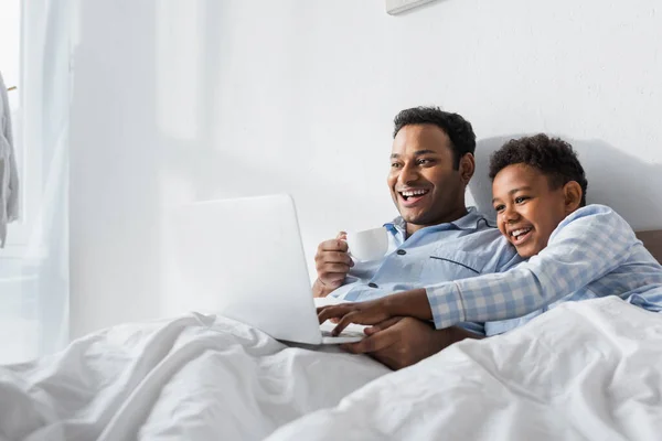 Animado afro-americano menino e pai assistindo comédia no laptop de manhã na cama — Fotografia de Stock
