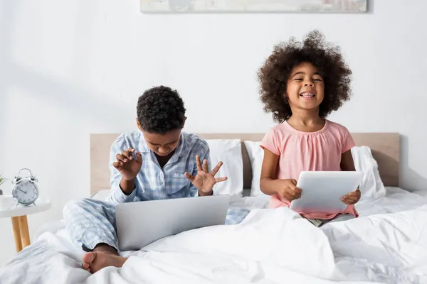 Africano americano chico usando laptop en cama cerca alegre hermana con digital tablet - foto de stock