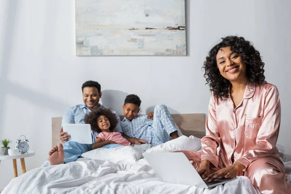 Happy african american woman working on laptop near blurred family with digital tablet in bed — Stock Photo