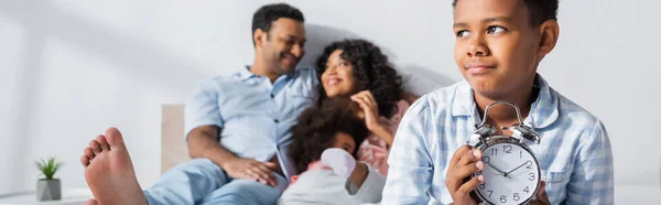 Découragé garçon afro-américain avec réveil près des parents flous et soeur dans la chambre, bannière — Photo de stock