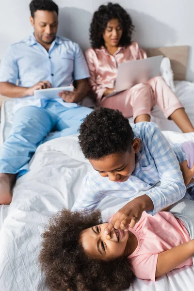 Alegre afroamericano hermanos divertirse en cama cerca borrosa padres con gadgets - foto de stock