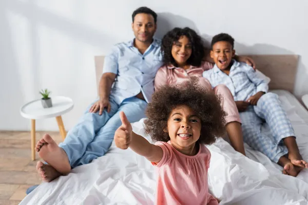 Cheerful african american girl showing thumb up at camera near blurred family in bedroom — Stock Photo