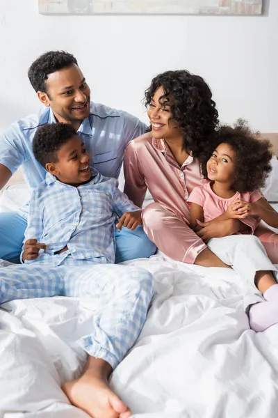 Sorrindo casal afro-americano abraçando crianças enquanto sentado na cama de manhã — Fotografia de Stock
