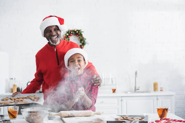 Uomo di mezza età con biscotti di Natale crudi vicino eccitato nipote afroamericano urlando vicino alla farina in cucina — Foto stock