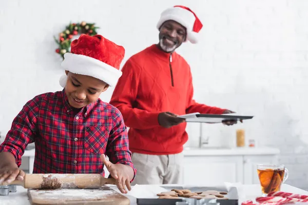 Homem americano africano desfocado com assadeira perto menino rolando massa enquanto prepara biscoitos de Natal — Fotografia de Stock
