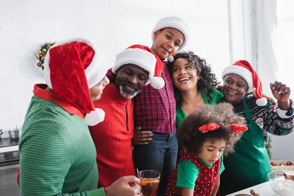 Веселая африканская американская семья в шляпах Санта-Клауса, обнимающая и улыбающаяся на кухне — стоковое фото