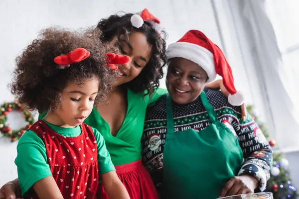 Pleased african american women in santa hats looking at girl in reindeer horns headband — Stock Photo