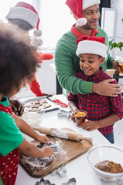 Homme afro-américain avec thé à la cannelle orange embrassant fils excité près de la famille préparant biscuits de Noël — Photo de stock