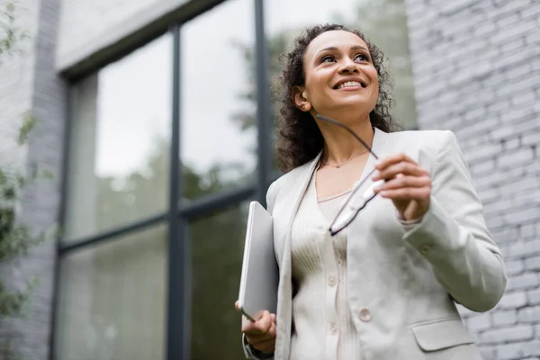 Vista a basso angolo di sorridente donna d'affari africana americana con laptop e occhiali vicino all'edificio sfocato — Foto stock