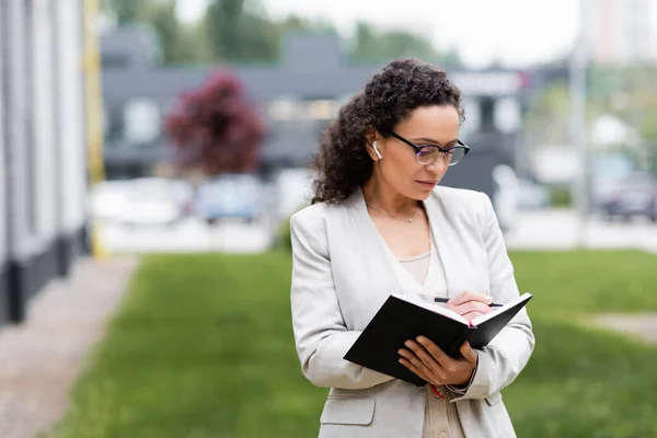 African american businesswoman in earphone and eyeglasses writing in notebook on urban street — Stock Photo