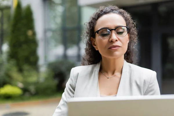 Afro-americana donna d'affari in occhiali da vista utilizzando il computer portatile offuscata sulla strada della città — Foto stock
