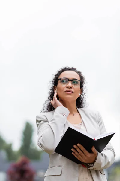 Vista de ángulo bajo de la mujer de negocios afroamericana sosteniendo portátil y ajustando auriculares al aire libre - foto de stock