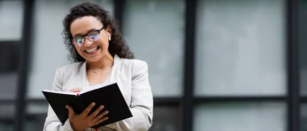 Femme d'affaires afro-américaine joyeuse dans les lunettes et l'écriture d'écouteurs dans le carnet à l'extérieur, bannière — Photo de stock