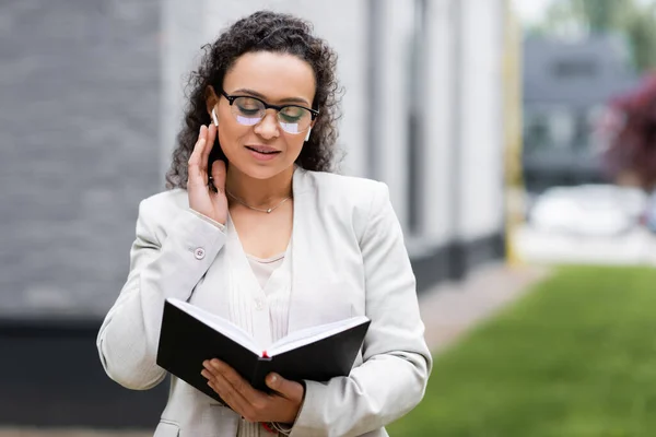 Mujer de negocios afroamericana tocando auriculares mientras mira en el cuaderno al aire libre - foto de stock