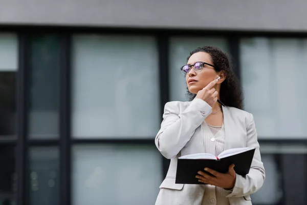 Afro-americana donna d'affari con notebook guardando altrove mentre la regolazione auricolare sulla strada della città — Foto stock