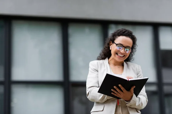 Alegre mujer de negocios afroamericana en anteojos y escritura de auriculares en cuaderno en la calle de la ciudad - foto de stock