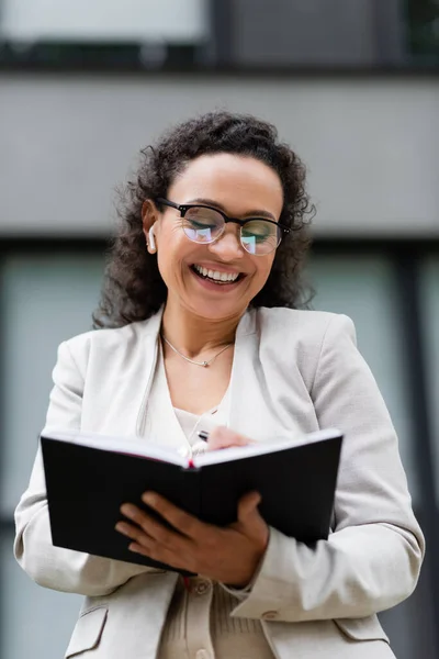 Exitosa mujer de negocios afroamericana en el auricular sonriendo mientras escribe en el cuaderno al aire libre - foto de stock