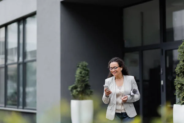 Afro-américaine femme d'affaires avec des papiers roulés souriant tout en utilisant un téléphone portable au premier plan flou à l'extérieur — Photo de stock