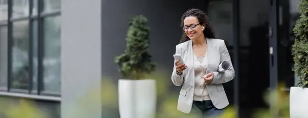 Heureux gestionnaire afro-américain avec des documents roulés en utilisant smartphone à l'extérieur sur le premier plan flou, bannière — Photo de stock