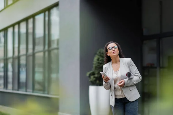 African american manager with cellphone and rolled papers looking away near blurred building — Stock Photo