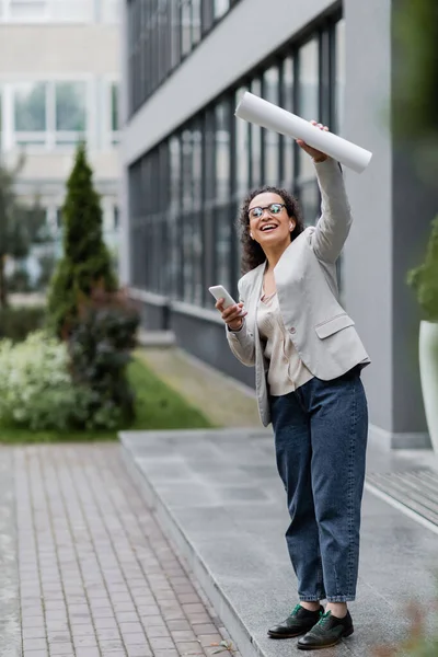 Alegre mujer de negocios afroamericana con teléfono móvil mirando hacia otro lado y agitando papeles enrollados cerca de edificio borroso - foto de stock