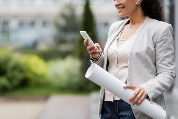 Cropped view of smiling african american businesswoman with rolled papers using smartphone outdoors — Stock Photo