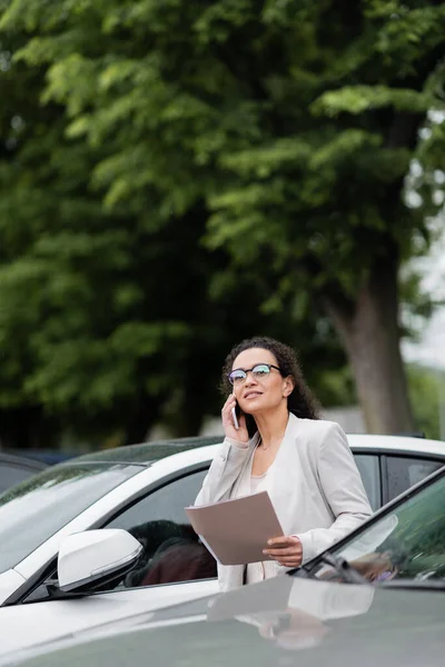African american businesswoman with paper folder looking away while calling on cellphone on car parking — Stock Photo