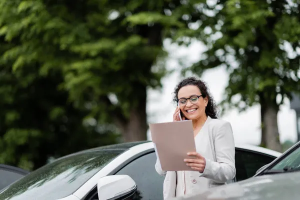 Mulher de negócios americana africana alegre falando no telefone celular enquanto olha para documentos no estacionamento do carro — Fotografia de Stock