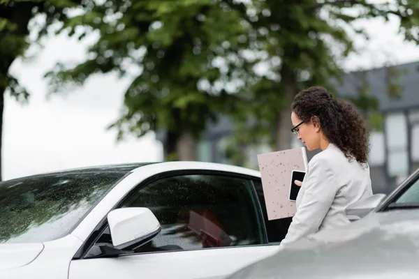 Mujer de negocios afroamericana con carpeta de papel y teléfono móvil cerca de coche en el estacionamiento - foto de stock
