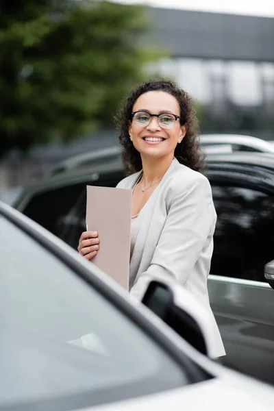 Donna d'affari afro-americana di successo con cartella di carta sorridente vicino a auto offuscate all'aperto — Foto stock