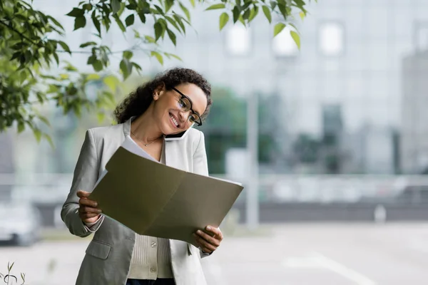 Femme d'affaires afro-américaine souriante en lunettes regardant des documents et parlant sur smartphone à l'extérieur — Photo de stock