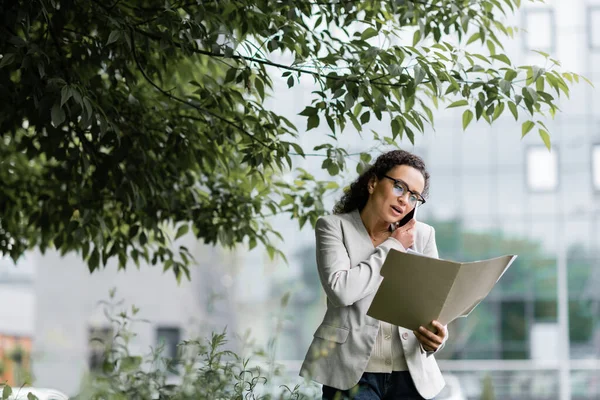 Mujer de negocios afroamericana mirando documentos durante la conversación en el teléfono inteligente al aire libre - foto de stock