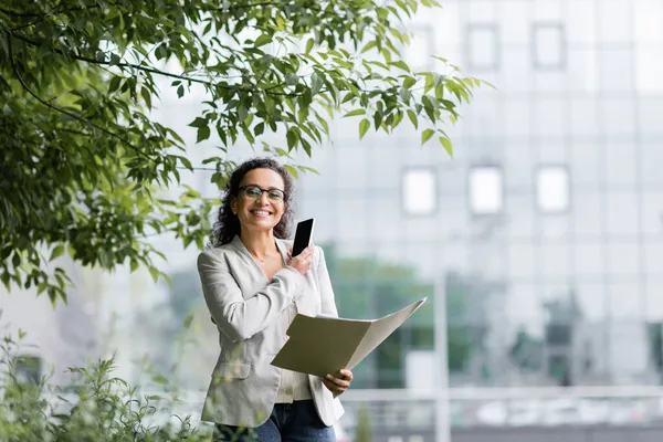 African american businesswoman with smartphone and paper folder smiling at camera under tree branches outdoors — Stock Photo