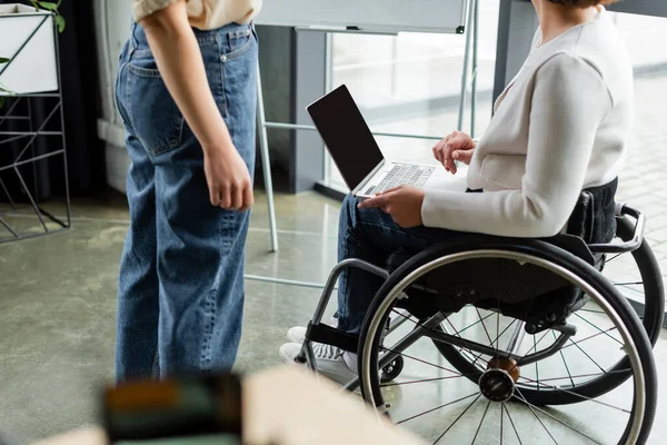 Vista cortada de empresária em cadeira de rodas segurando laptop com tela em branco perto colega no escritório — Fotografia de Stock