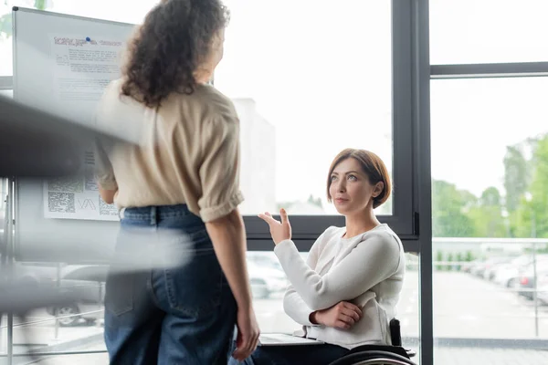 Rückseite der afrikanisch-amerikanischen Geschäftsfrau nahe Kollegin im Rollstuhl zeigt auf Flipchart im Büro — Stockfoto