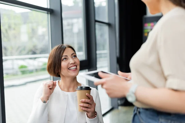 Emocionada empresaria sosteniendo café para llevar mientras muestra gesto de victoria cerca borrosa colega afroamericano con tableta digital - foto de stock