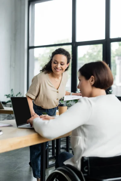 Smiling african american businesswoman looking at laptop near blurred colleague with disability in office — Stock Photo