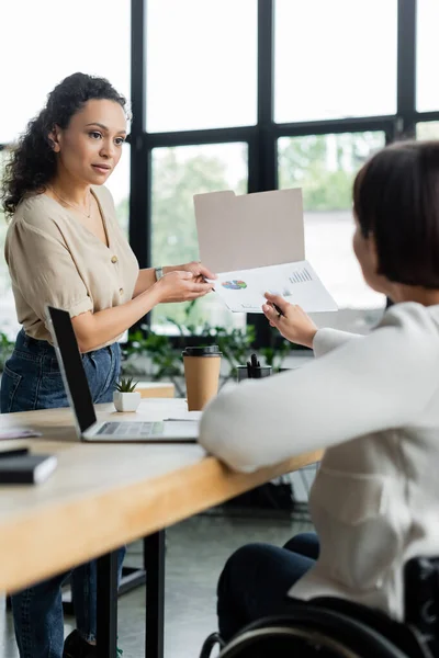 African american businesswoman and her colleague with disability pointing at documents with graphs in office — Stock Photo