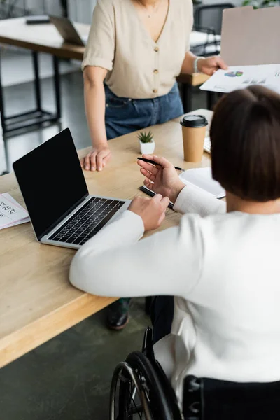 Vista posteriore della donna d'affari con disabilità che punta al computer portatile vicino collega afro-americano con documenti — Foto stock