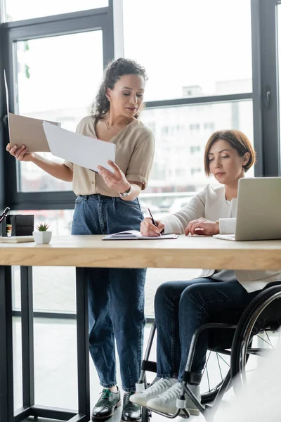 Donna d'affari afroamericana in possesso di documenti vicino collega in sedia a rotelle scrittura in notebook — Foto stock